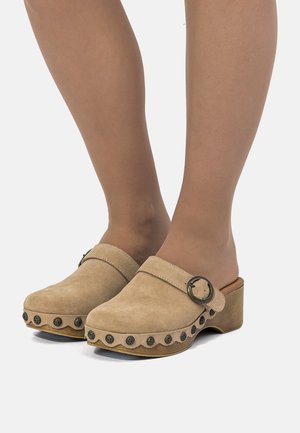 Zoccoli in camoscio beige con tacchi in legno e borchie nere decorative, indossati con calze velate color nudo su piedi, su sfondo bianco.