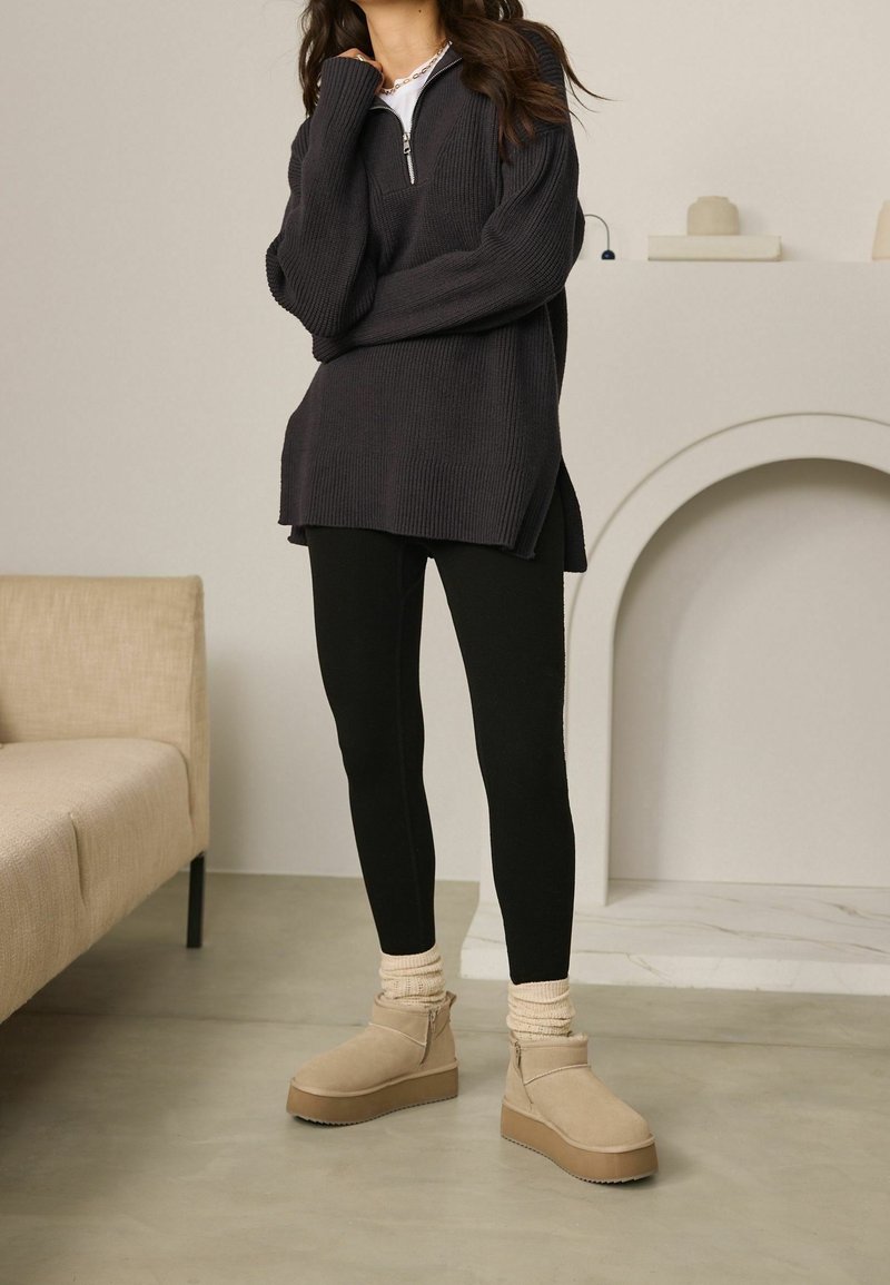 Maglione a coste grigio scuro con colletto zip, abbinato a leggings neri e stivali alla caviglia beige con fusti testurizzati e suole a platform.