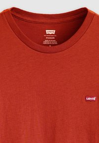 Cămașă T-shirt roșie din bumbac, cu guler rotund, având un mic logo Levi's pe partea stângă și o etichetă de întreținere în interiorul gulerului.