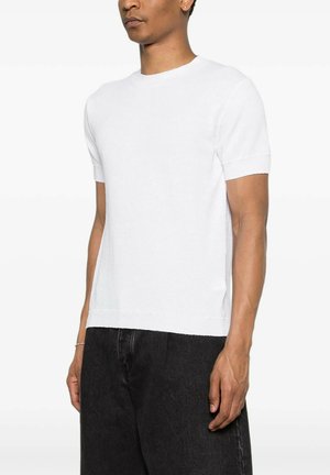 Homme portant un pull blanc uni à manches courtes et un pantalon noir, debout devant un fond blanc uni.