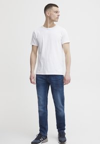 Camiseta de algodón blanca combinada con jeans de mezclilla azules. El modelo está de lado; la prenda tiene un corte clásico y un ajuste casual. Zapatillas texturizadas completan el look.