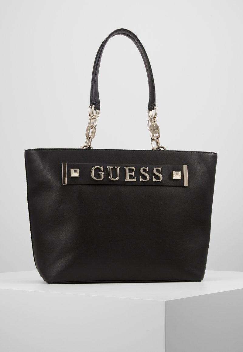 Sac cabas noir GUESS avec poignées en chaîne dorée et lettrage décoratif doré sur surface en cuir lisse.