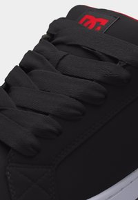 Κοντινή λήψη μαύρου αθλητικού παπουτσιού με χοντρές κορδόνια, κόκκινο και μαύρο λογότυπο DC στη γλώσσα και λευκή σόλα ορατή στο κάτω άκρο.