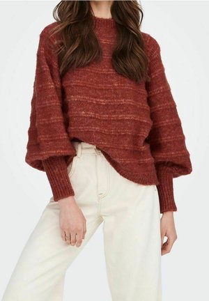 Femme portant un pull ample et texturé couleur rouille avec des manches bouffantes, associé à un pantalon taille haute blanc cassé, sur un fond uni.