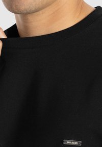 Schwarzes Baumwoll-T-Shirt mit Rundhalsausschnitt und feinen Rippendetails, das ein kleines metallisches Etikett mit dem Text "SNILODOX" zeigt.