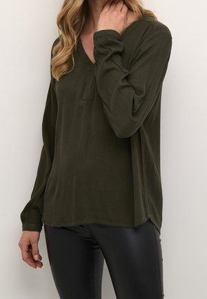 Femme portant un chemisier texturé manches longues couleur olive foncé avec un col en V et un pantalon en cuir noir, la main près du cou, sur un fond clair uni.