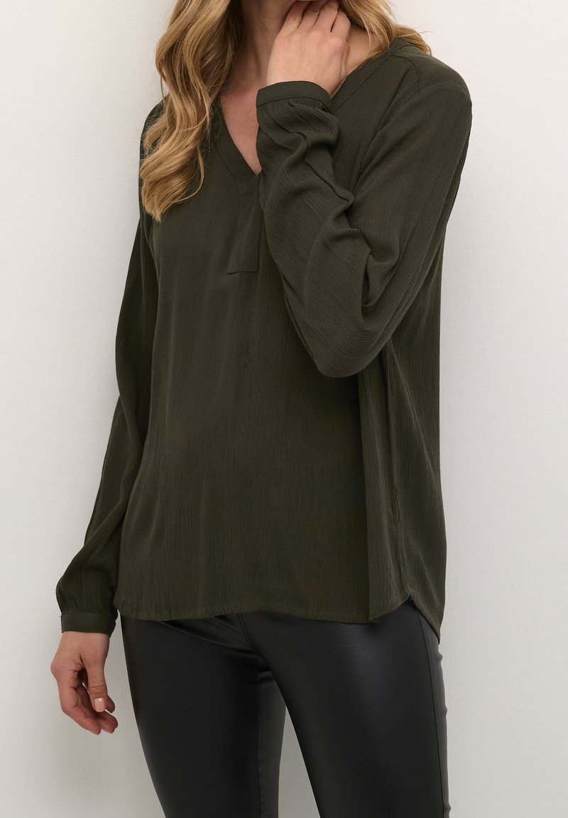 Femme portant un chemisier texturé manches longues couleur olive foncé avec un col en V et un pantalon en cuir noir, la main près du cou, sur un fond clair uni.