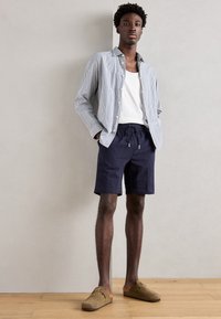 Gestreepte overhemd met knopen, witte tanktop en marineblauwe shorts met trekkoord; draagt beige instappers; staat tegen een neutrale achtergrond.