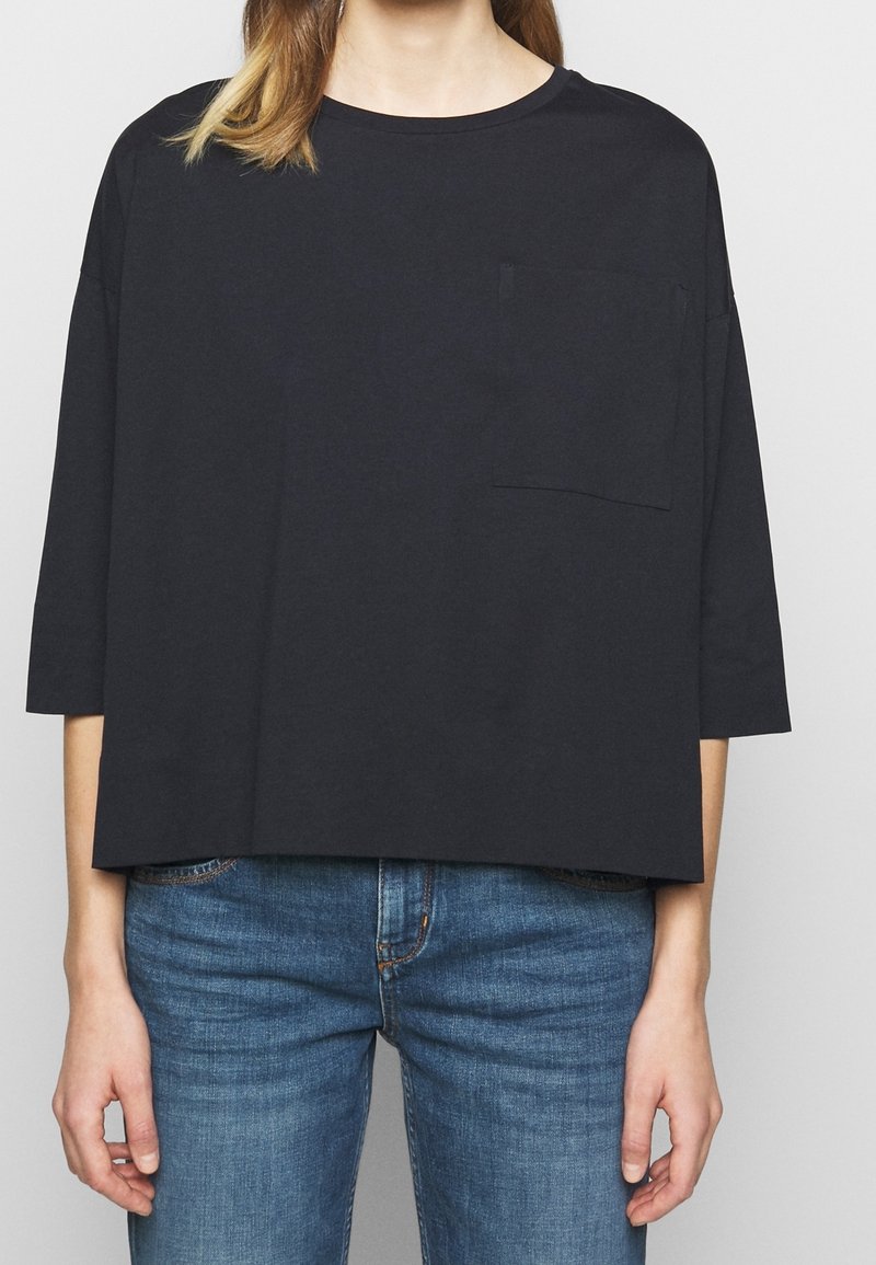Schwarzes, kurzes T-Shirt mit rundem Halsausschnitt, drei Viertelärmeln und kleiner Brusttasche. Kombiniert mit blauen Jeans. Glatte Textur.