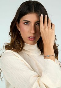 Bracciale d'argento con una pietra blu centrale, anelli circolari, indossato su un polso contro una maglietta chiara e testurizzata. Sfondo neutro.
