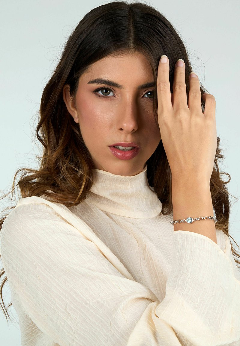 Bracciale d'argento con una pietra blu centrale, anelli circolari, indossato su un polso contro una maglietta chiara e testurizzata. Sfondo neutro.