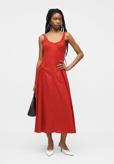 Γυναίκα με μακριά πλεξούδα που φοράει αμάνικο κόκκινο midi φόρεμα, κρατώντας μια σκούρα τσάντα και άσπρες μυτερές γόβες σε απλό φόντο.