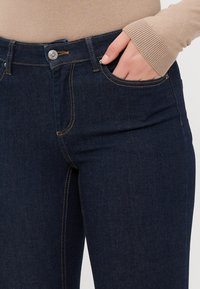 Tmavě modré džíny vyrobené z denimu, s knoflíkovým zapínáním, kapsami a kontrastním žlutým prošíváním podél švů.
