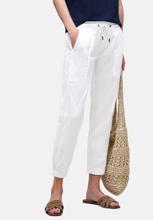 Person iført hvide cropped bukser med snøre, navy bluse, bærer en beige flettet taske og brune slip-on sandaler.