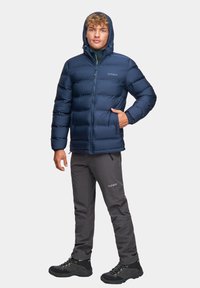 Granatowa kurtka puchowa z kapturem, wyposażona w zamek błyskawiczny i elastyczne mankiety; zestawiona z szarymi spodniami outdoorowymi i czarnymi butami wędrówkowymi.
