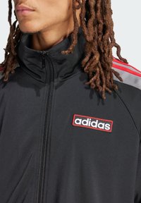 Veste de survêtement Adidas noire avec un col montant, fermeture éclair, design à trois bandes rouges sur les épaules et un logo brodé rouge et blanc.