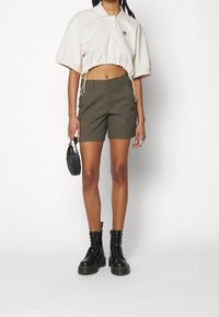 Beige Poloshirt mit gekürztem Schnitt und Kordelzugsaum, kombiniert mit olivgrünen Shorts mit Logo und schwarzen Schnürstiefeln in Knöchelhöhe. Hält eine kleine schwarze Tasche.