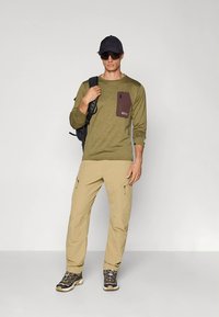 Olívazöld hosszú ujjú ing barna zsebel, bézs katonai nadrággal és fekete túracipővel párosítva. A modell fekete hátizsákot visel.