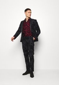 Svart blomstmønstret dress med en mørkerød skjorte under. Designet med slim fit og teksturert stoff. Kombinert med mørke sko.