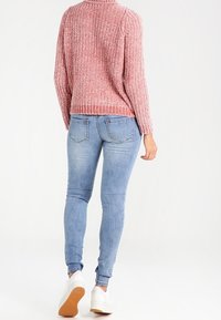 Suéter de punto rosa con cuello redondo, combinado con jeans ajustados azul claro. Zapatillas blancas con una suela texturizada visible completan el conjunto.