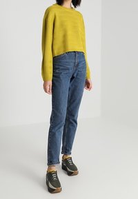 Un pull jaune court, un jean taille haute bleu et des baskets vert foncé avec des accents jaunes et des lacets blancs. Design texturé simple.