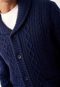 Cardigan navy con colletto a scialle, caratterizzato da un motivo a treccia e costine, bottoni neri e una finitura in maglia testurizzata.