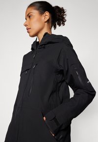 Black waterproof jacket featuring a zippered front, dual chest pockets, and textured fabric. Equipped with a hood and adjustable cuffs.