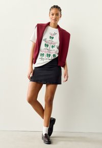 T-shirt bianca con motivo grafico verde e rosa, cardigan senza maniche color bordeaux, gonna in denim nera e mocassini scuri con calzini bianchi.