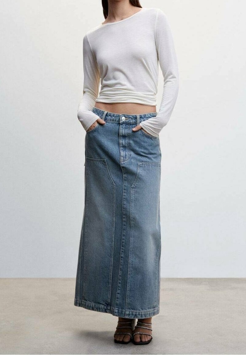 Femme portant un haut blanc à manches longues, une jupe maxi en jean taille haute bleu clair avec de grandes poches avant, et des sandales noires à brides.