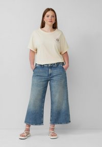 Cremfarbiges, übergroßes T-Shirt mit umgeschlagenen Ärmeln und Sonnenmotiv; hellblaue weit geschnittene Jeans; weiße Riemchensandalen; neutraler Hintergrund.