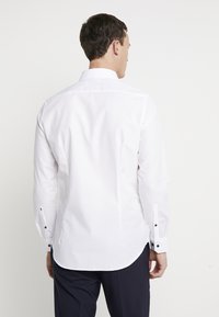 Camicia bianca a maniche lunghe con colletto classico, orlo curvato e bottoni blu scuro sui polsini. Tessuto liscio e leggero con una vestibilità su misura.