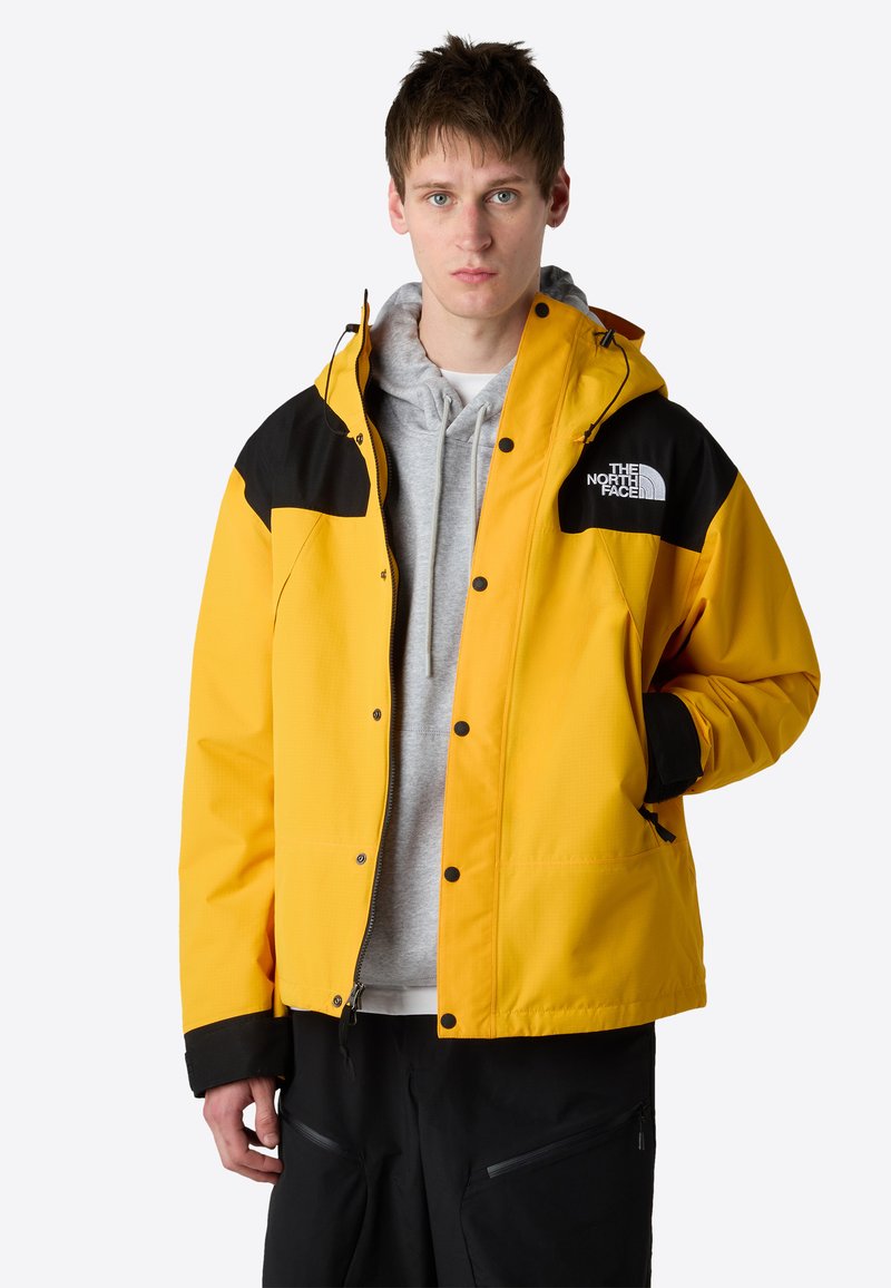 Žlutá voděodolná bunda s černými detaily, předním zipem a kapucí. Obsahuje otvory na kapsy a logo na levé hrudi. Texturovaná tkanina.