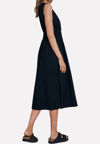 Robe noire sans manches avec une taille cintrée, une jupe fluide et une fente sur le côté ; associée à des sandales foncées avec une semelle en liège.