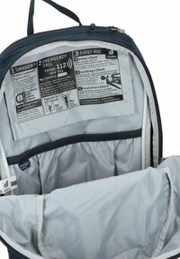 Interior de una mochila con un forro de nailon gris. Incluye instrucciones de emergencia impresas en texto negro sobre un fondo gris. Presenta múltiples compartimentos.