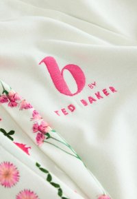 Witte stof met roze bloemdessins aan de rand. Heeft "b by TED BAKER" geborduurd in roze schrift op het hoofdgedeelte.