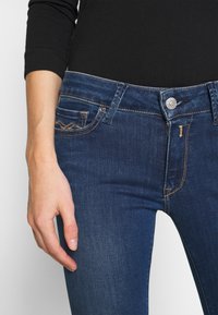 Jeans de mezclilla azul oscuro con un diseño ajustado, que cuentan con pespuntes en dorado y un cierre de botón, resaltados por un detalle en el bolsillo.