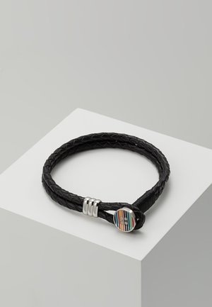 Schwarzes geflochtenes Lederarmband mit silbernem Verschluss und einem runden, mehrfarbig gestreiften Knopfdetail, präsentiert auf einem weißen, geometrischen Sockel.