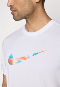 Hvit bomull t-skjorte med et stort, flerfarget Nike swoosh-logo i oransje, blå og ferskenfarger. Korte ermer og rund hals.