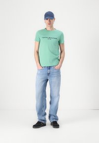 Green cotton t-shirt with black logo, distressed light blue jeans, and black shoes. Accessories include a blue cap. Simple, casual look.