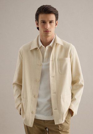 Giovane uomo con una giacca beige abbottonata sopra una polo bianca e pantaloni kaki, in piedi con le mani in tasca davanti a uno sfondo semplice.