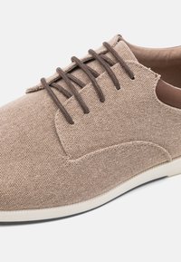 Zapato de lona beige con un suave acento de cuero marrón, que presenta seis cordones marrones y una puntera redondeada. Superficie texturizada con una suela ligera.