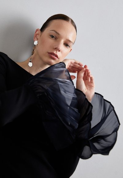 Black top with sheer, ruffled sleeves in a layered tulle design. Model wearing circular earrings. Neutral background. Subtle texture visible.