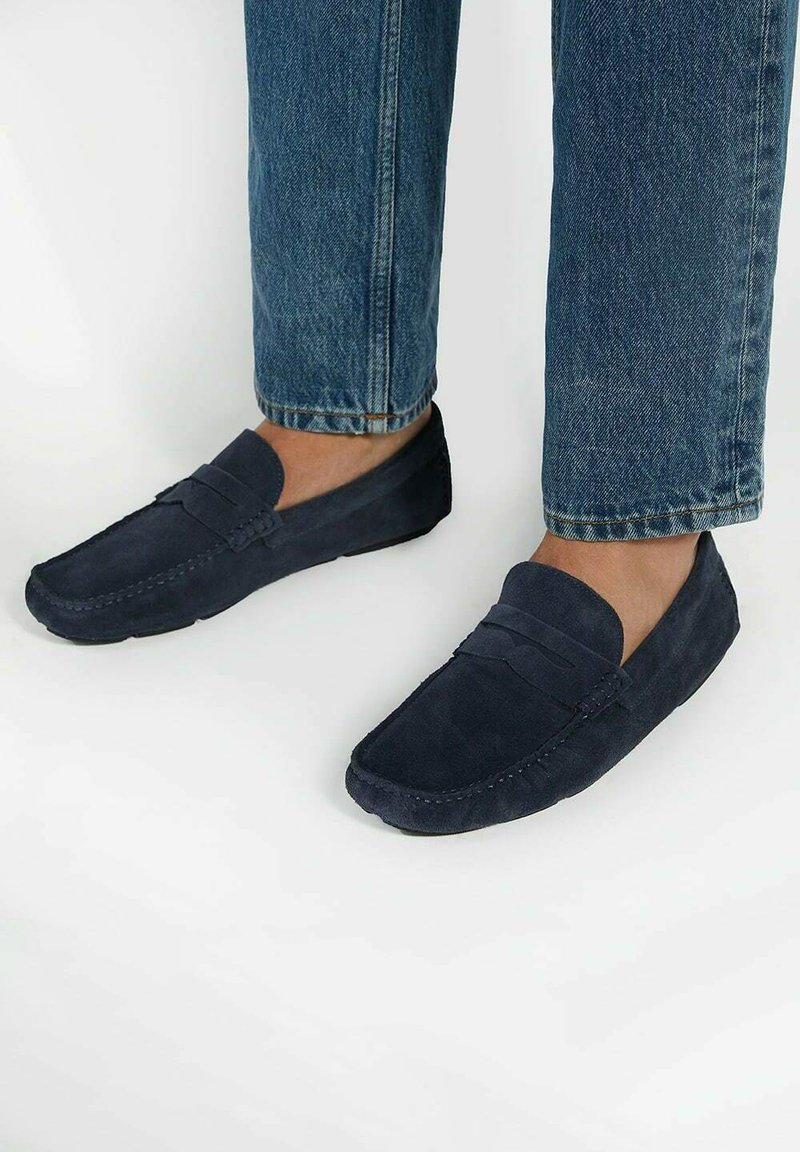 Navy blue suede loafers with a flat sole, featuring a stitched design and slip-on style. Worn with light blue denim pants.