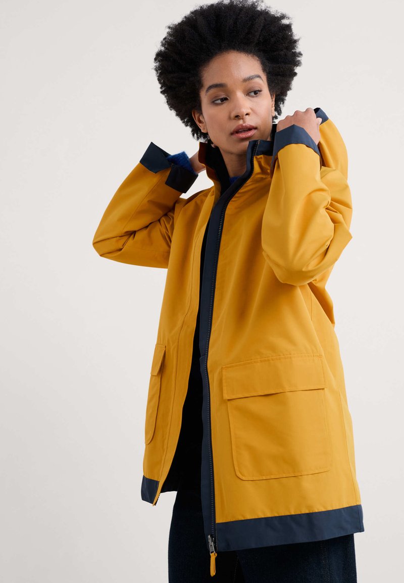 Geltona nepralaidi striukė su aukštu apykakle, tamsiai mėlynais rankogaliais ir dviem priekinėmis kišenėmis; turi užtrauktuką ir laisvą siluetą.