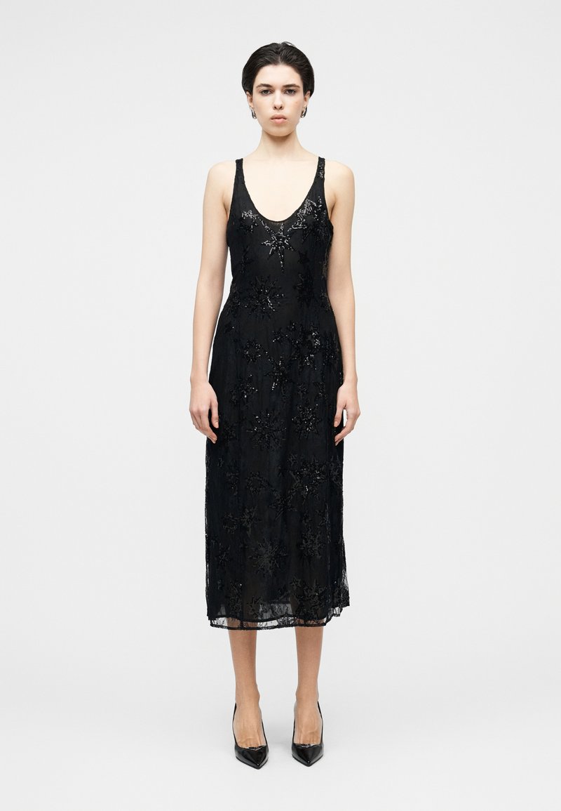 Robe en dentelle noire avec un col en V, un voile semi-transparent à motifs floraux, des détails perlés et un ourlet à la hauteur du genou. Chaussures �à talons hauts.