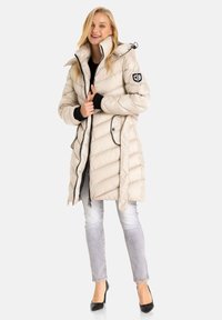 Beige Daunenjacke mit Kapuze, Reißverschluss, seitlichen Taschen, gerippten schwarzen Bündchen, gestepptem Muster und einem Logo-Patch am linken Arm.