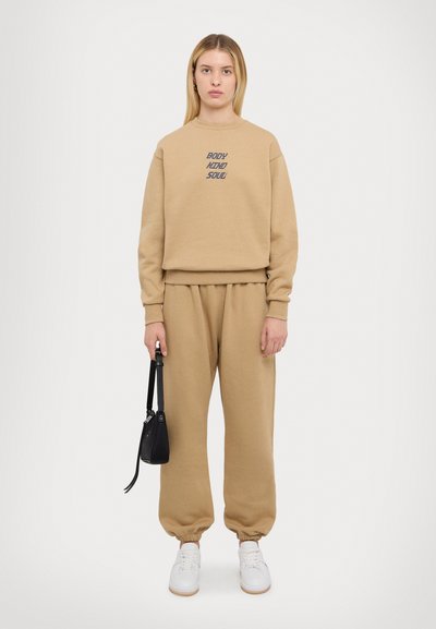 Beige sweatshirt en bijpassende joggers van zachte stof. De sweatshirt heeft de tekst "BODY MIND SOUL". Witte sneakers en een kleine zwarte tas.