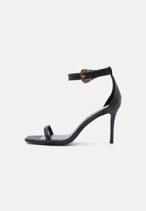 Black high-heeled sandal with thin ankle strap featuring an ornate gold buckle and a single toe strap, textured leather finish.