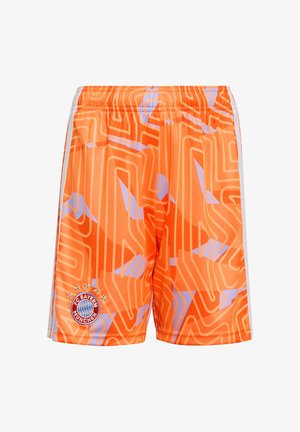Un niño pequeño está de pie con las manos detrás de la espalda, vistiendo un uniforme de fútbol del Bayern de Múnich de color naranja brillante con botas negras sobre un fondo blanco.