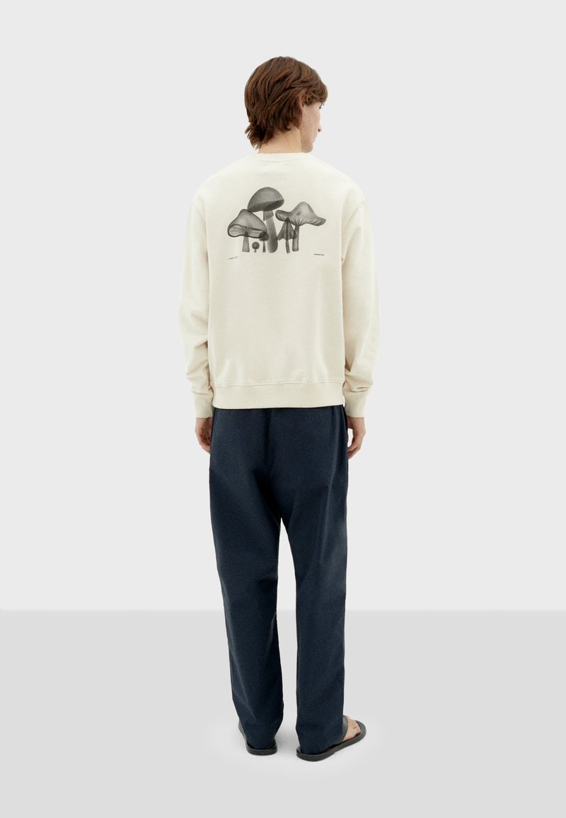 Crème-kleurige sweatshirt met een monochrome paddenstoelprint op de achterkant, gecombineerd met donkere broeken en casual schoeisel.